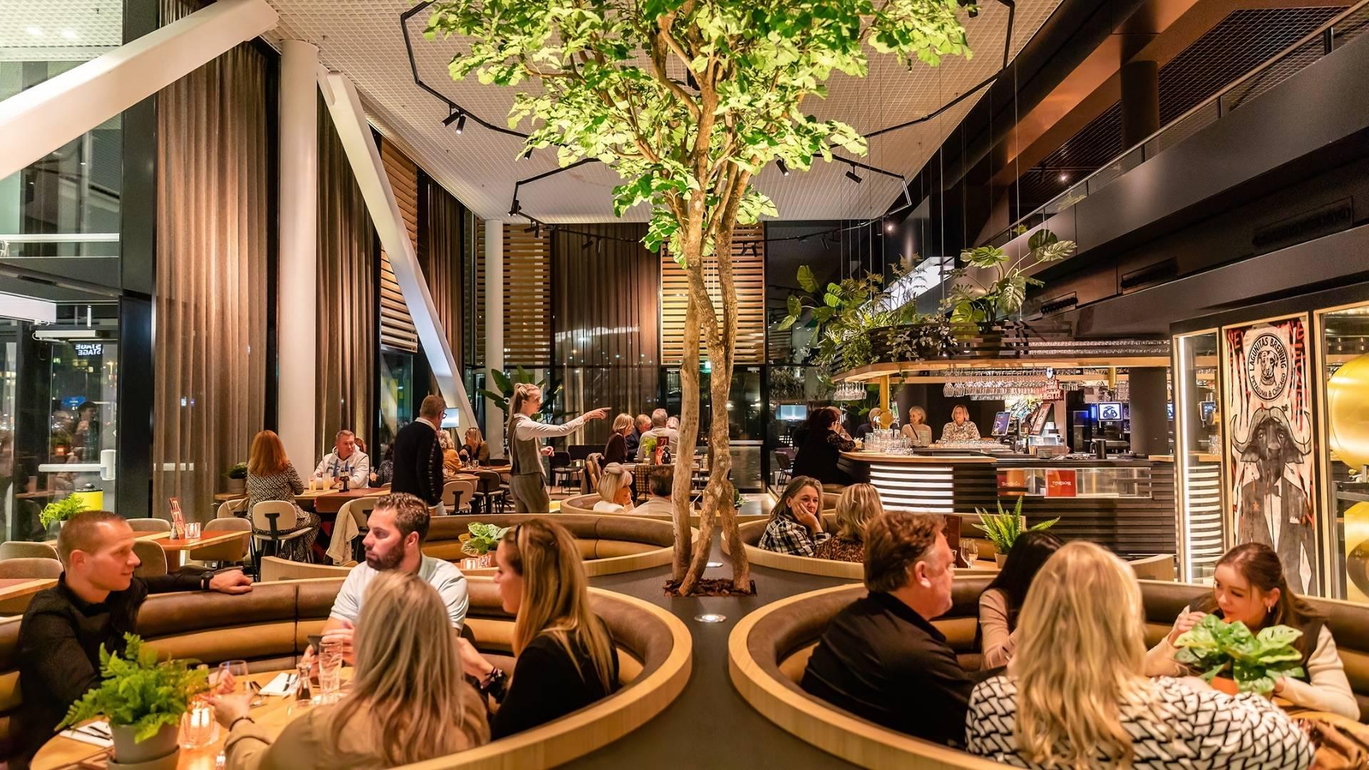 Druk bezet restaurant met ronde zitjes en een boom in het midden